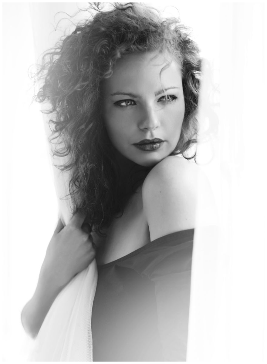 Černobílá boudoir momentka ženy u okna zamotané v záclonách s odhalenými rameny