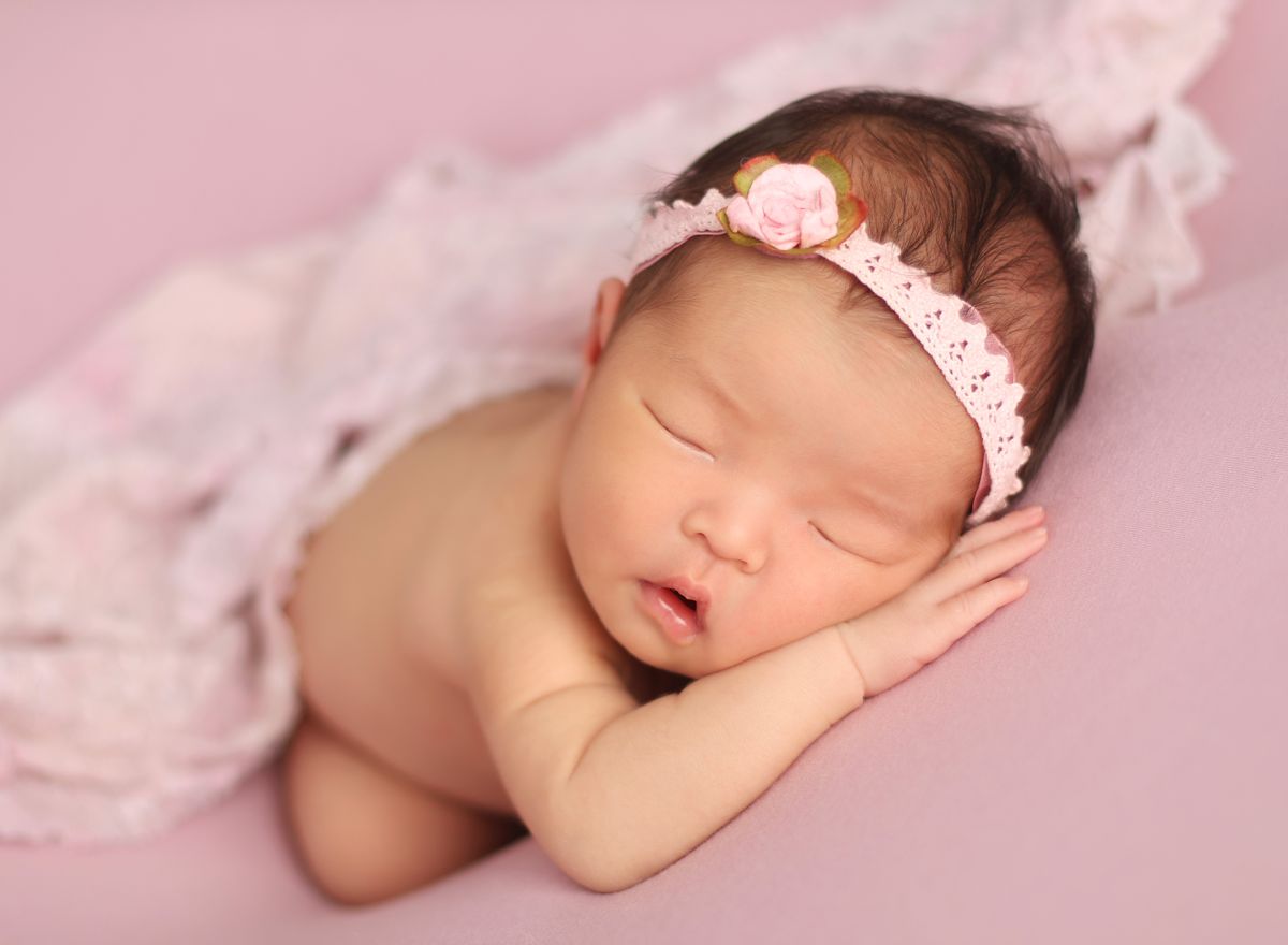 Jemná fotografie spinkajícího novorozeňátka s čelenkou na růžovém podkladě v denním světle 